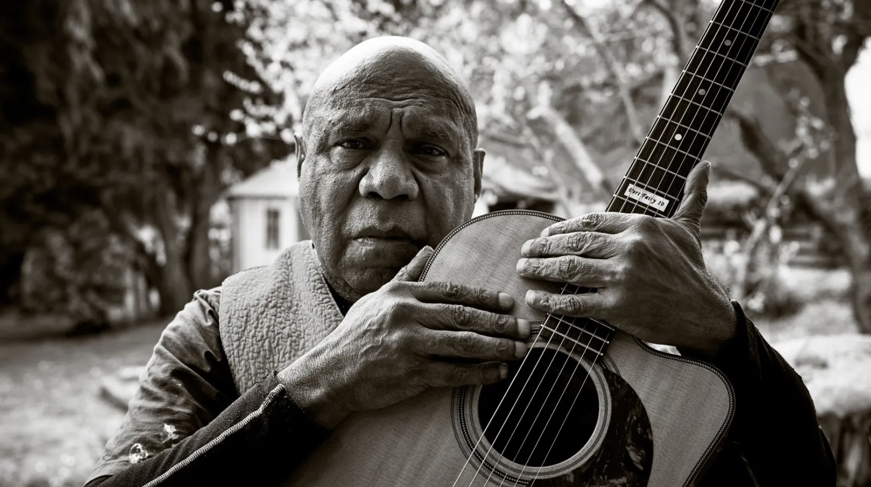 Black and white headshot of an older man holding a guitar with a house and trees in the background