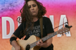 Woman with long brown hair and closed eyes holding a guitar onstage in a t-shirt that reads WORKING CLASS HERO