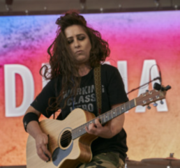 Woman with long brown hair and closed eyes holding a guitar onstage in a t-shirt that reads WORKING CLASS HERO