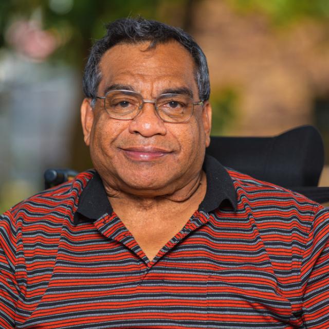 Headshot of a seated smiling man wearing glasses and a collared shirt with red and grey stripes