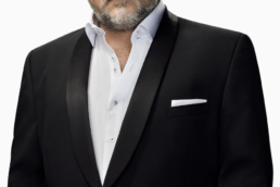 A grey-haired man with a beard wearing a black suit jacket