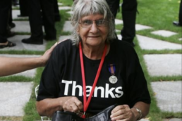 A seated woman with grey hair wearing a medal, a red lanyard and a black t-shirt that reads 