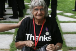 A seated woman with grey hair wearing a medal, a red lanyard and a black t-shirt that reads 
