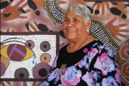 Smiling woman with short grey hair wearing a flowered blouse standing in front of a First Nations mural