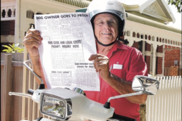 Elderly man wearing a red shirt and white helmet sitting on a motorbike holding up a newspaper