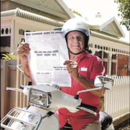 Elderly man wearing a red shirt and white helmet sitting on a motorbike holding up a newspaper