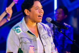 A black-haired woman wearing a jean jacket singing into a microphone on a stage