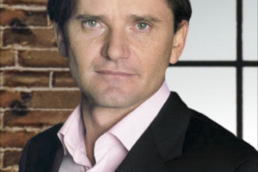 Headshot of a brown-haired man wearing a black suit jacket and white collared shirt against a brick wall with a window