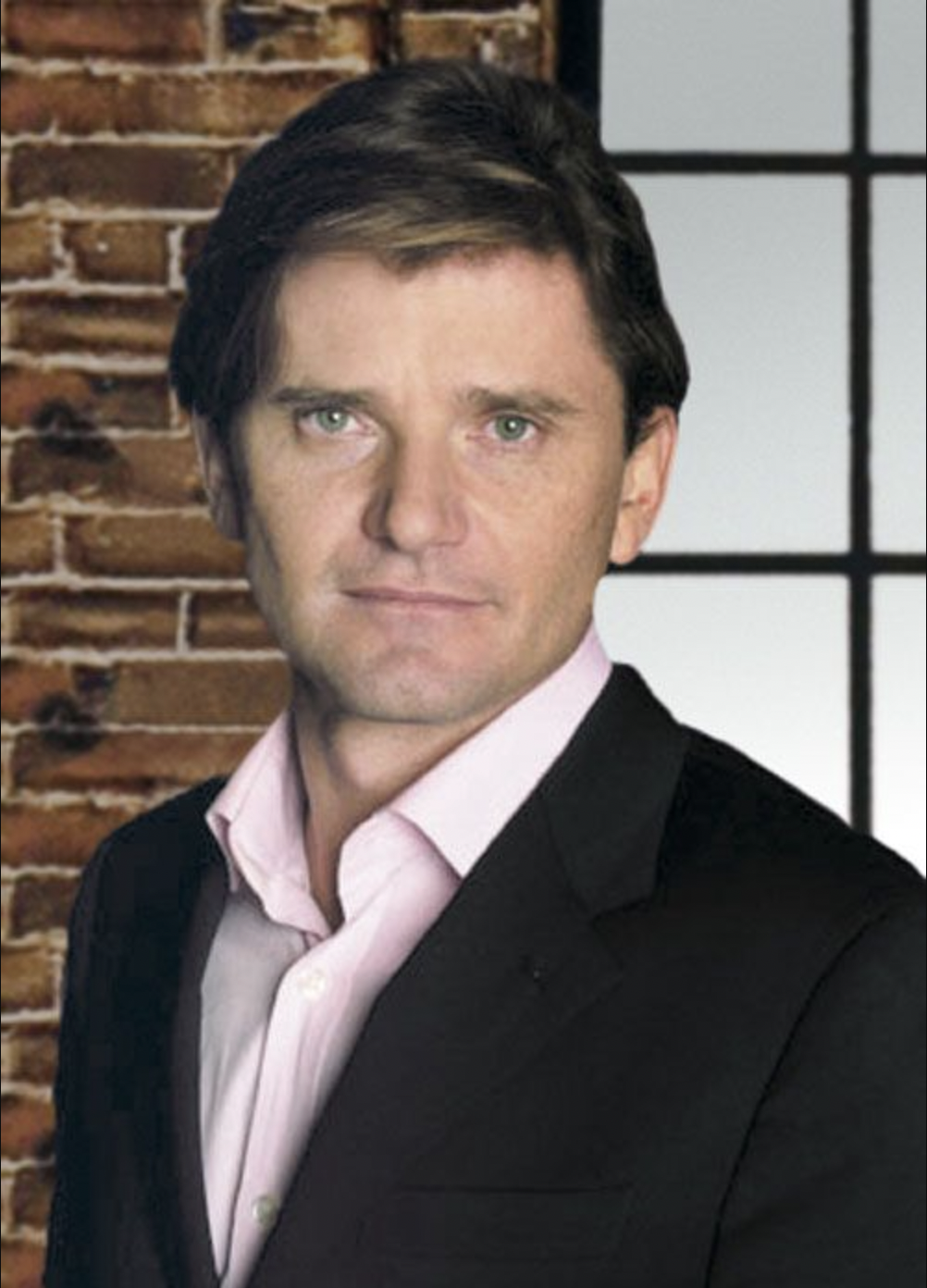 Headshot of a brown-haired man wearing a black suit jacket and white collared shirt against a brick wall with a window