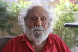Headshot of an elderly man with bushy grey hair and beard wearing a red polo shirt