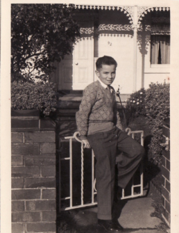Black and white photo of a young boy wearing a jumper andleaning on a gate in front of a house