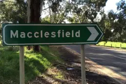 Green street sign with the word Macclesfield against a background of a tree lined road.