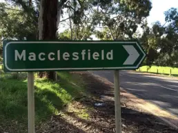 Green street sign with the word Macclesfield against a background of a tree lined road.