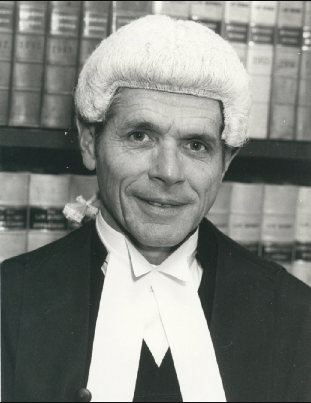 Black and white headshot of a smiling man wearing a High court judge wig and robes against a background of textbooks