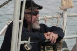 Grey haired man with a beard wearing a black hat, shirt, and sunglasses standing on a boat looking at the water