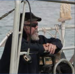 Grey haired man with a beard wearing a black hat, shirt, and sunglasses standing on a boat looking at the water