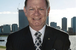 Headshot of a smiling man wearing a black suit with matching tie and handkerchief with a river and city skyline backdroop