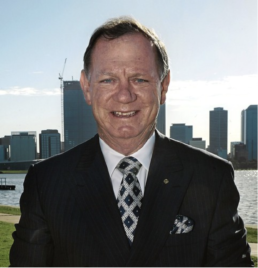Headshot of a smiling man wearing a black suit with matching tie and handkerchief with a river and city skyline backdroop