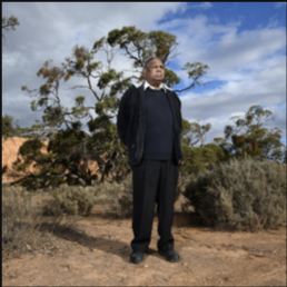 An older man dressed in black pants, jacket, and coat standing outdoors on red soil with bushland in the background