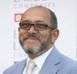 Headshot of a smiling man with a greying beard and mustache wearing glasses and a light blue suit and tie