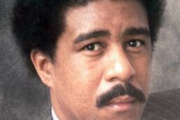 Headshot of a man with a mustache and afro wearing a grey suit and tie.