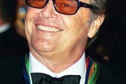 Headshot of an older smiling man wearing red sunglasses and a suit and bowtie