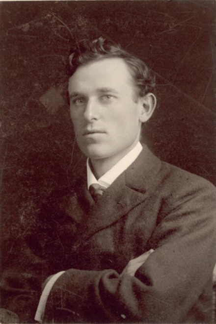 Black and white headshot of a man wearing an old fashioned suit and jacket with short curly hair with a neutral expression