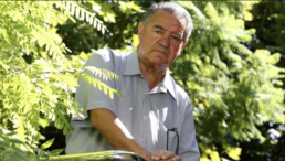 An older man with grey hair and a neutral expression standing at angle surrounded by tree branches