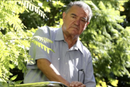 An older man with grey hair and a neutral expression standing at angle surrounded by tree branches