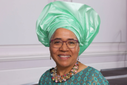 Headshot of a smiling woman with glasses wearing a brightly colored necklace, turquoise top and traditional Gele headscarf
