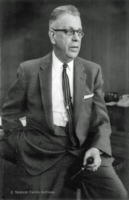Black and white image of an older man with glasses standing with one foot propped up wearing a suit and tie
