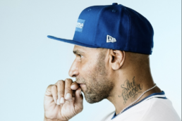 Profile of a bearded man with a neck tattoo wearing a gold chain, white t-shirt, and blue baseball cap with his hand touching his face