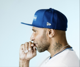 Profile of a bearded man with a neck tattoo wearing a gold chain, white t-shirt, and blue baseball cap with his hand touching his face