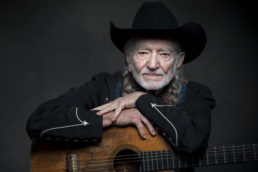 Gray-haired man with long pigtails and a beard wearing a black cowboy hat and resting his hands on top of an acoustic guitar