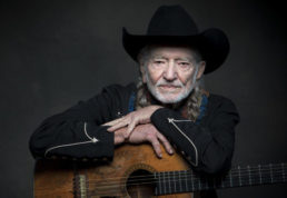 Gray-haired man with long pigtails and a beard wearing a black cowboy hat and resting his hands on top of an acoustic guitar