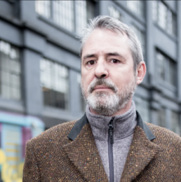 Headshot of a greyhaired man with a beard and moustache wearing a brown tweed coat with a building in the background