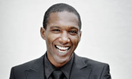 Headshot of a short haired man with a big smile wearing a black shirt, tie, and black suit jacket against a white backdrop