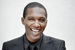 Headshot of a short haired man with a big smile wearing a black shirt, tie, and black suit jacket against a white backdrop