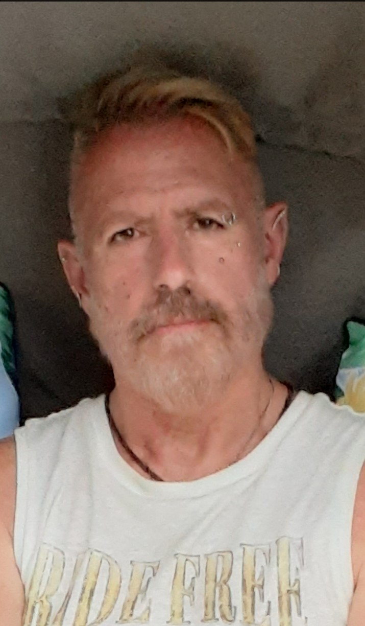 Headshot of a middle-aged man with ginger coloured moustache and an eyebrow ring wearing a white t-shirt and black necklace