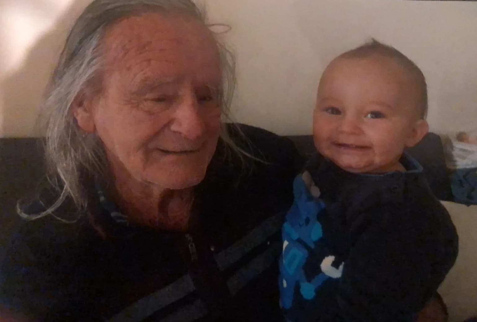 An elderly man with long grey hair holding a smiling baby in a blue shirt