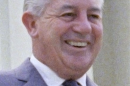 Headshot of a smiling grey haired man wearing a grey suit and tie with a white building in the background