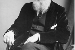 Black and white image of a seated man with white hair, glasses, and a bushy beard wearing an old fashioned suit and waistcoat