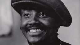 Black and white close up of a smiling man with a mustache wearing a beret and turtleneck.