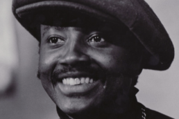 Black and white close up of a smiling man with a mustache wearing a beret and turtleneck.