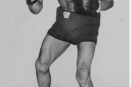 Black and white image of a muscular, shirtless man with an Afro wearing boxing clubs in a fighting stance