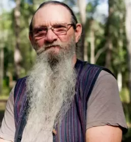Headshot of a balding with a long grey beard wearing glasses and a grey t-shirt with a blue vest against a background of a forest.