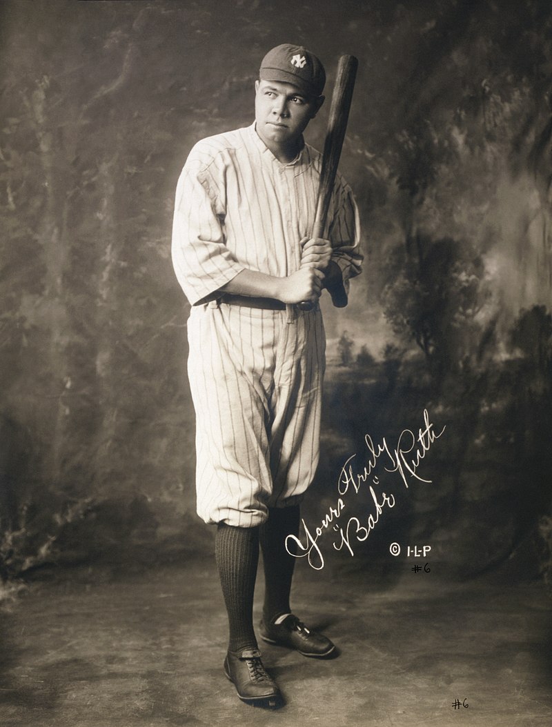 Black and white image of a man wearing a white baseball uniform, cap, and black shoes with knee high black socks posed holding a baseball bat with Babe Ruth's autograph on the corner of the image
