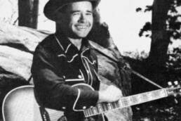 Black and white image of a smiling man in a cowboy hat holding a guitar in front of a fallen tree