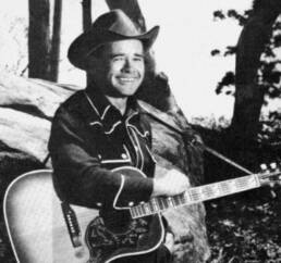 Black and white image of a smiling man in a cowboy hat holding a guitar in front of a fallen tree