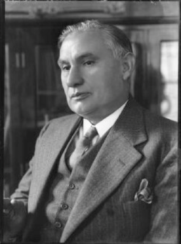 Black and white headshot of a grey haired man with a somber expression wearing a suit, jacket, and tie with a bookshelf in the background